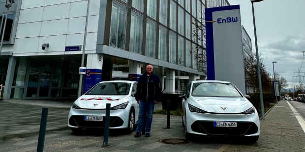 Two white cupra born vehicles parked outside an enbw, office building, with a fleet manager standing between the cars on a cloudy day.