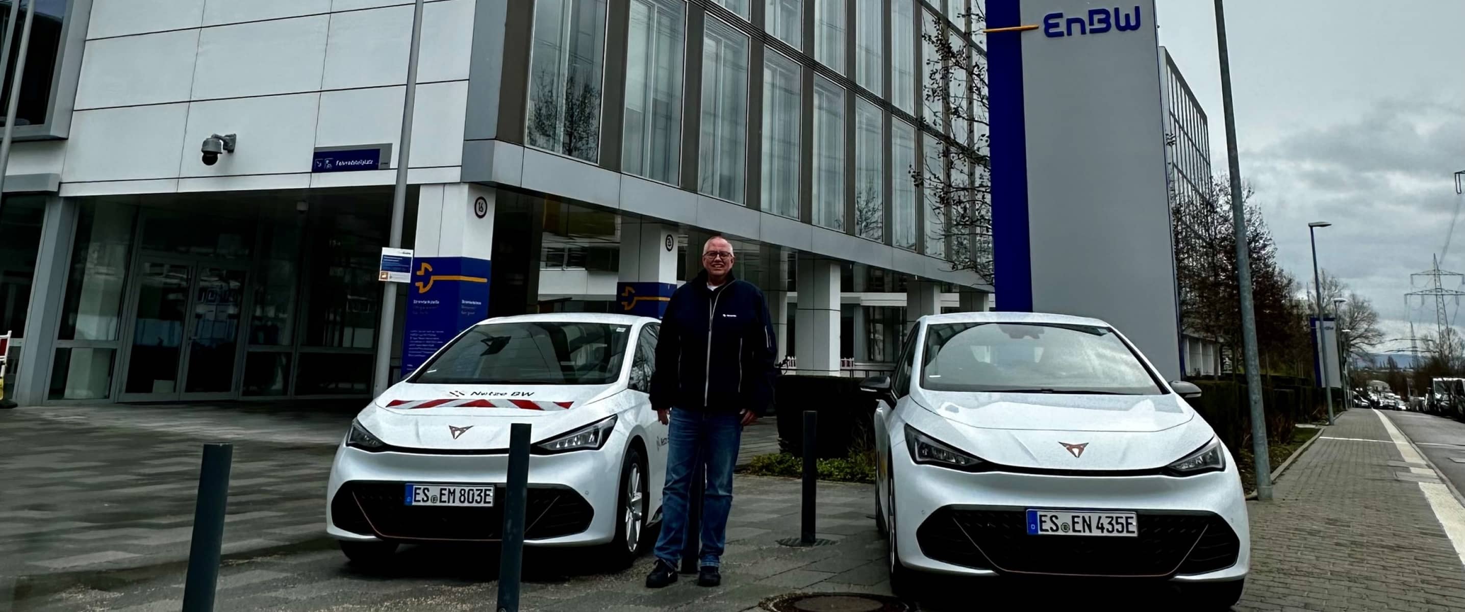 Two white cupra born vehicles parked outside an enbw, office building, with a fleet manager standing between the cars on a cloudy day.