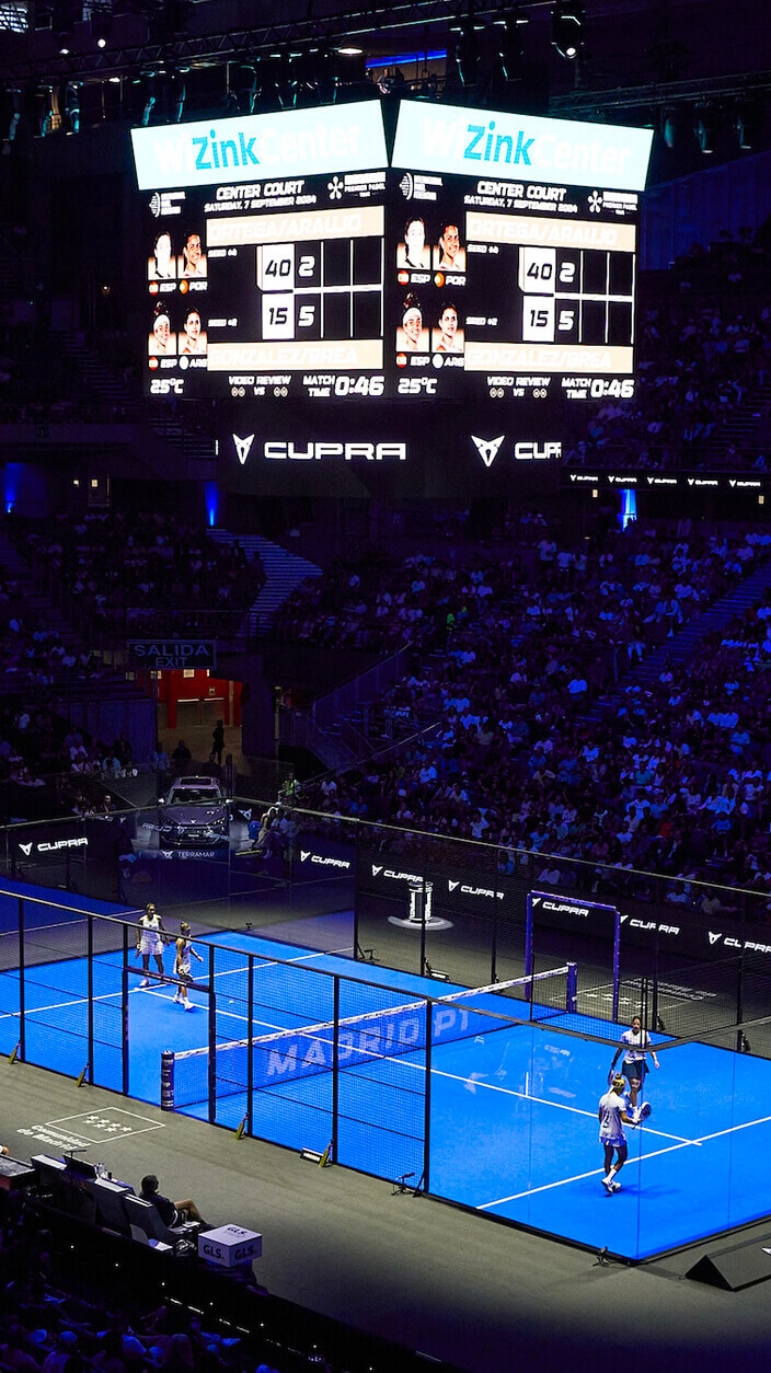 A padel court illuminated by blue lights, seen from above during a game within a large stadium filled with spectators. Two pairs of players are on the court and a large scoreboard is visible in the background.